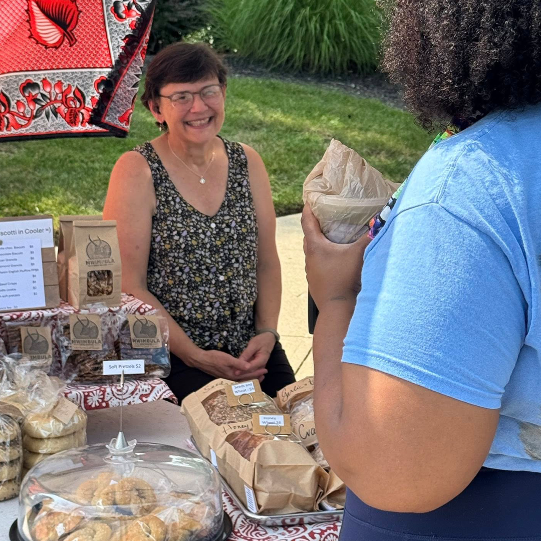 Smiling vendor sells baked goods at an outdoor market booth.